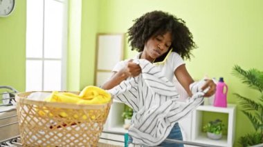 African american woman talking on smartphone hanging clothes on clothesline at laundry room