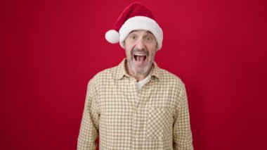 Orta yaşlı bir adam, kırmızı arka planda sürpriz bir ifadeyle Noel şapkası takıyor.