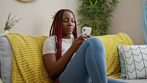 快乐的非洲裔美国女人 辫子享受舒适的家 坐在沙发上 用智能手机开心地在网上发短信 脸上挂着灿烂的笑容 — 图库视频影像
