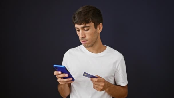 若いヒスパニック系男性は オンラインで買い物をしたり クレジットカードを持ったり スマートフォンでタイピングしたりしている 黒い壁の背景に対して隔離されて 投資の問題に直面している強調された大人の男 — ストック動画