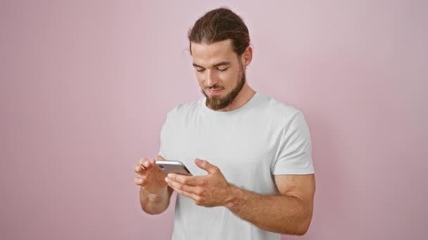 年轻的惊慌失措的男人用智能手机对着孤立的粉色背景微笑 — 图库视频影像