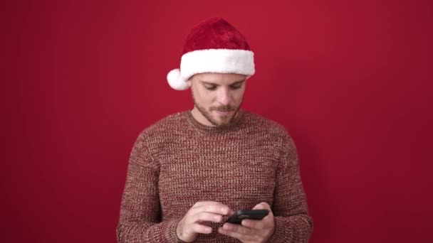 単離された赤い背景の上にクリスマスの帽子を身に着けている勝者表現のスマートフォンを使用する若い白人男性 — ストック動画