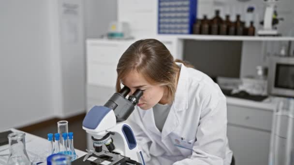 年轻自信的金发女科学家 两臂交叉地微笑着 坐在实验室桌旁 用显微镜在繁忙的科学实验室里研究医学 — 图库视频影像