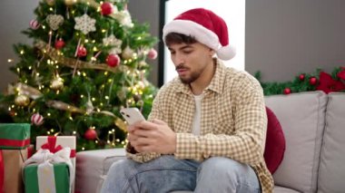 Genç Arap adam akıllı telefon kullanıyor. Yılbaşı ağacının yanında oturuyor. Evde üzgün üzgün bakıyor.