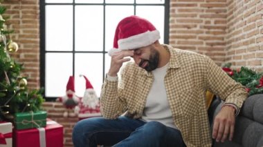 Noel şapkası takan genç İspanyol adam evde stresli bir şekilde oturuyor.