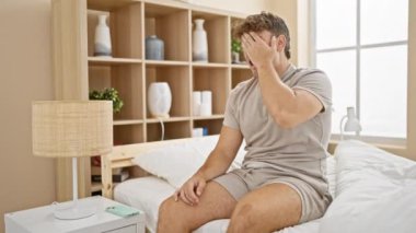 Yorgun, esneyen genç İspanyol bir adam, akıllı telefon kullanarak yatakta oturuyor, uzun bir gece boyunca yatak odasında mesajlaşmaktan yorgun..