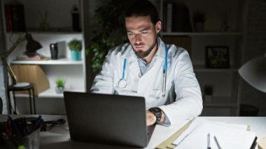 Genç İspanyol doktor, masada dizüstü bilgisayar kullanıyor. Klinikte kollarını kavuşturup el kol hareketi yapıyor.