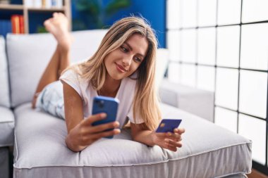 Young blonde woman using smartphone and credit card lying on sofa at home