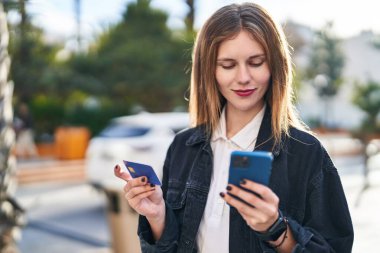Genç sarışın kadın parkta akıllı telefon ve kredi kartıyla alışveriş yapıyor.