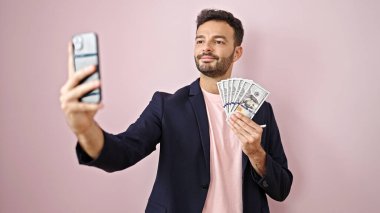 Genç İspanyol iş adamı dolar tutuyor ve izole edilmiş pembe arka plan üzerinden akıllı telefondan selfie çekiyor.