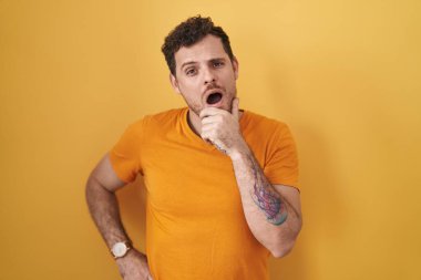 Sarı arka planda duran İspanyol genç adam, elleri çenesinde, inanamama, şaşkınlık ve şaşkınlık içinde bakıyor. 