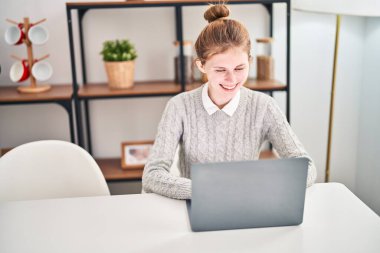 Genç sarışın kadın yemek odasındaki masada dizüstü bilgisayar kullanıyor.