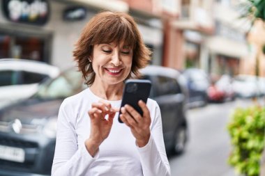 Sokakta akıllı telefon kullanan orta yaşlı bir kadın gülümsüyor.
