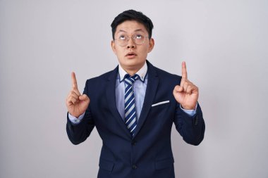 Genç Asyalı takım elbiseli ve kravatlı adam şaşırmış ve şaşırmış bir şekilde yukarı bakıyor ve parmaklarıyla işaret ediyor ve kollarını kaldırıyor.. 