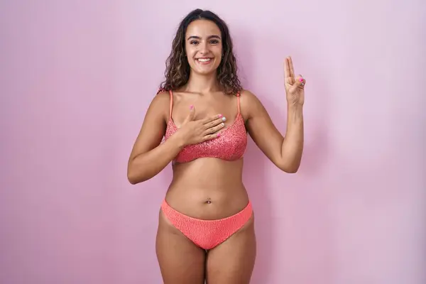 若いヒスパニック女性は 胸と指の手で汗をかいた笑顔のピンクの背景の上にランジェリーを着て 忠誠心の誓いを作ります — ストック写真
