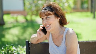 Orta yaşlı bir kadın gülümsüyor. Parktaki akıllı telefondan konuşuyor.