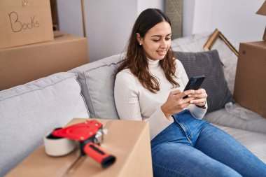 Young beautiful hispanic woman smiling confident using smartphone at new home