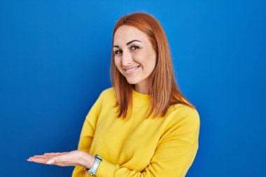 Young woman standing over blue background inviting to enter smiling natural with open hand 