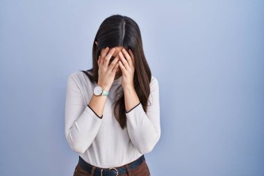 Mavi arka planda duran genç esmer kadın. Ağlarken suratını ellerle kapatan üzgün bir ifade. depresyon kavramı. 