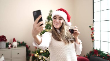 Genç ve güzel İspanyol kadın akıllı telefonuyla video görüşmesi yapıyor. Yılbaşı ağacının yanında duruyor.