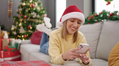 Evdeki Noel ağacının yanındaki koltukta akıllı telefon kullanan genç sarışın kadın.