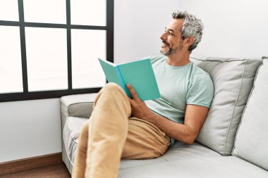Orta yaşlı, kır saçlı, kitap okuyan adam evdeki kanepede oturuyor.