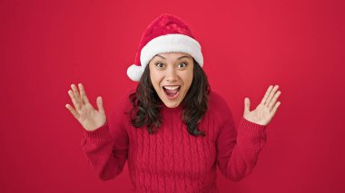 Genç, güzel, İspanyol bir kadın sürpriz bir ifadeyle izole edilmiş kırmızı arka planda noel şapkası takıyor.