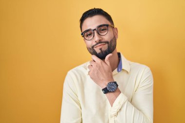 İş kıyafetleri ve gözlük takan İspanyol genç adam kameraya kendinden emin bir şekilde bakıyor kollarını kavuşturup, çenesini kaldırarak gülümsüyor. Olumlu düşünmek. 