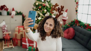 Orta yaşlı İspanyol kadın akıllı telefonuyla selfie çekiyor. Evdeki yılbaşı ağacının yanında oturuyor.