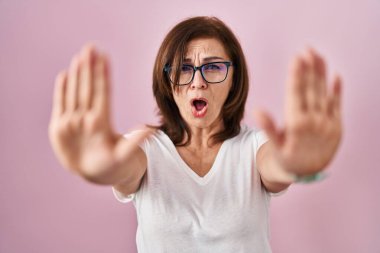 Middle age hispanic woman standing over pink background doing stop gesture with hands palms, angry and frustration expression 