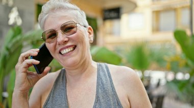 Orta yaşlı, kır saçlı, gülümseyen, kendine güvenen bir kadın parkta akıllı telefondan konuşuyor.