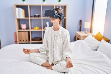 Young caucasian woman doing yoga exercise using virtual reality glasses at bedroom