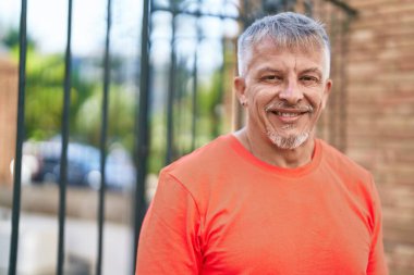 Orta yaşlı, kır saçlı, gülümseyen, kendine güvenen bir adam sokakta duruyor.