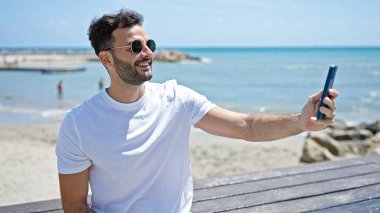 Young hispanic man smiling confident making selfie by the smartphone at seaside