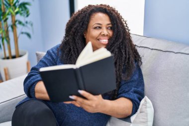 Evdeki kanepede oturan Afro-Amerikalı kadın kitap okuyor.