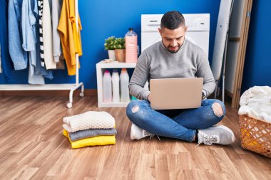 Young latin man using laptop waiting for washing machine at laundry room