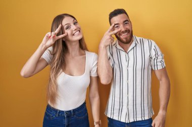 Young couple standing over yellow background doing peace symbol with fingers over face, smiling cheerful showing victory 