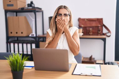 Young blonde woman working at the office wearing glasses shocked covering mouth with hands for mistake. secret concept.  clipart