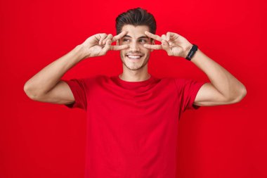 Kırmızı arka planda dikilen İspanyol genç adam barış sembolü yapıyor. Yüzünde parmakları, gülümseyerek zaferi gösteriyor. 