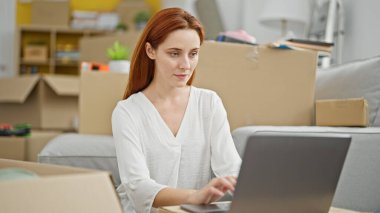 Genç kızıl saçlı kadın yeni evinde dizüstü bilgisayar kullanıyor.