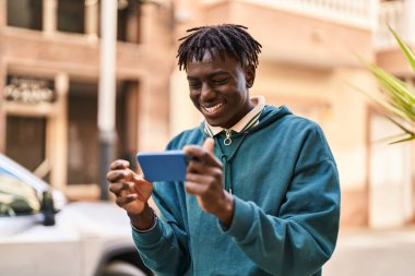 Afro-Amerikalı adam gülümsüyor. Sokakta akıllı telefondan videolar izliyor.