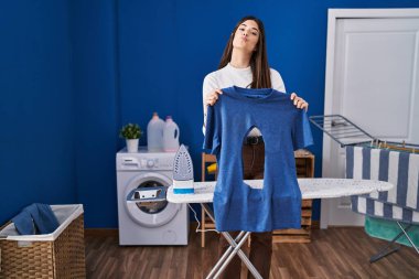 Çamaşır odasında ütülü ütülü, elinde demir gömleği olan genç bir kadın kameraya bakıp öpüşürken çok güzel ve seksi görünüyor. aşk ifadesi. 