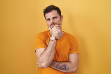 Sarı arka planda duran Latin asıllı genç adam kameraya kendinden emin bir şekilde bakıyor kolları çapraz ve çenesi yukarıda gülümsüyor. Olumlu düşünmek. 