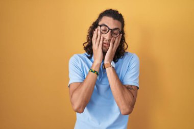 Young hispanic man standing over yellow background tired hands covering face, depression and sadness, upset and irritated for problem 