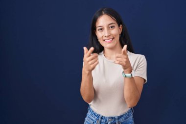 Mavi arka planda dikilen genç İspanyol kadın mutlu ve komik yüzlü parmaklarını kameraya doğrultuyor. iyi enerji ve titreşimler. 