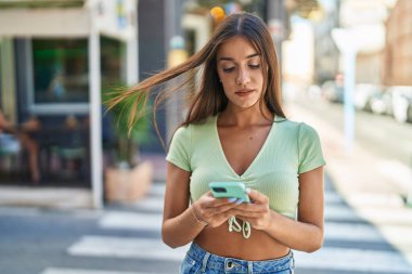 Genç, güzel, İspanyol bir kadın sokakta rahat bir ifadeyle akıllı telefon kullanıyor.