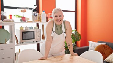 Orta yaşlı, gri saçlı, gülümseyen bir kadın yemek odasında masasında kendinden emin bir şekilde duruyor.