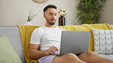 Genç Arap adam evdeki kanepede dizüstü bilgisayar kullanıyor.