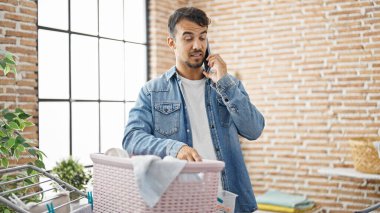 Genç İspanyol adam akıllı telefondan konuşuyor çamaşırhanede çamaşır ipine kıyafet asıyor.