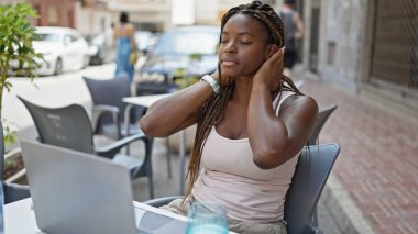 Afro-Amerikalı bir kadın dizüstü bilgisayar kullanıyor, saçlarını tarıyor kahve dükkanının terinde terliyor.